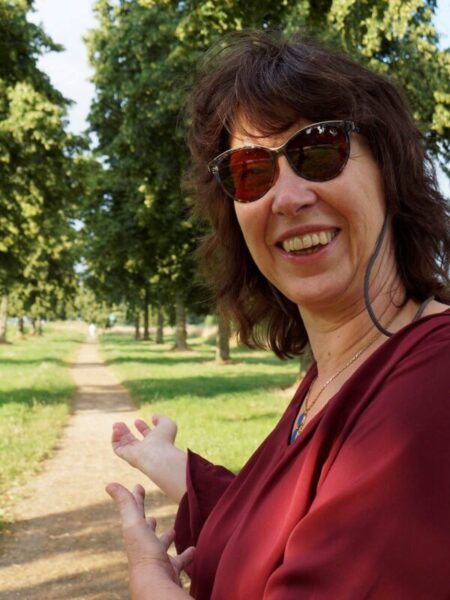 Foto von Anne Niesen, Seheldin. Draußen, mit Sonnenbrille. Sie lächelt herzlich und weißt auf einen Weg