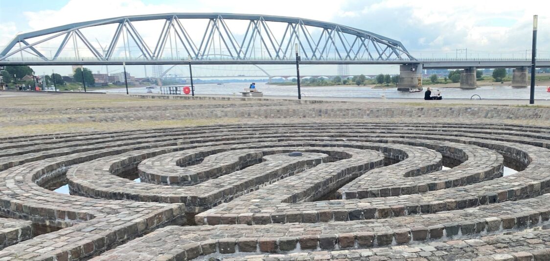 Das Foto zeigt ein Labyrinth aus Steinen und im Hintergrund eine Brücke, als Symbole für das Leben, die Suche, das Ankommen.